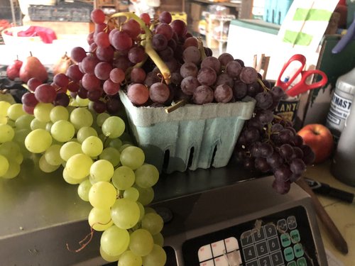 A basket of assorted grapes, including red and green varieties, displayed on a scale in a market setting, with additional fruits and tools visible in the background.