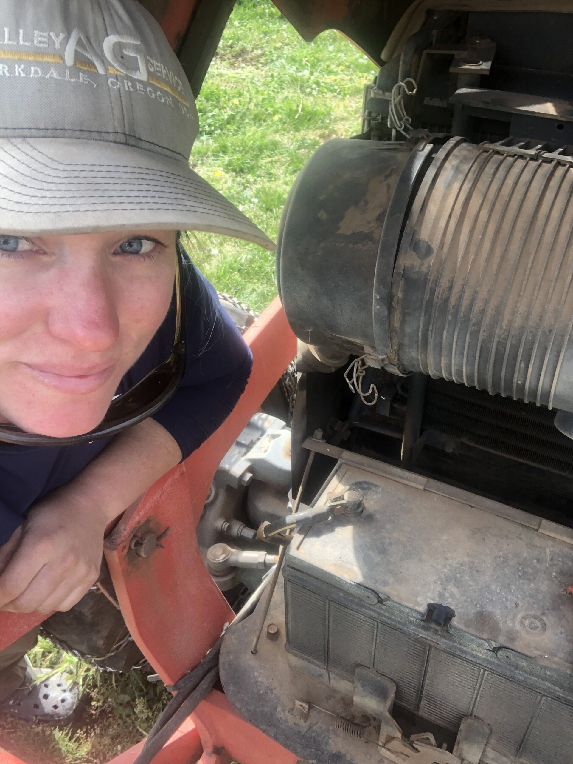 Woman inspecting a tractor engine, wearing a cap and sunglasses, surrounded by mechanical components and grass in the background.